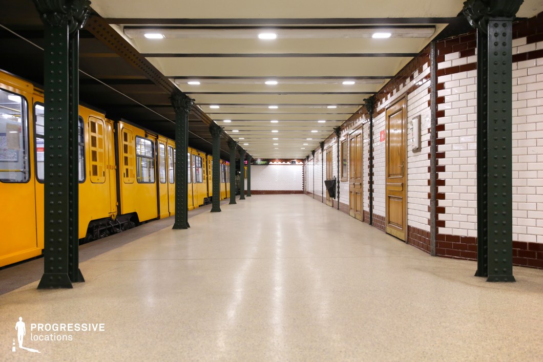 This image captures a vintage underground metro platform with a yellow train positioned alongside dark green riveted iron columns. The platform is framed by classic white subway tiles accented with red trim, while wooden doors and paneling add warmth to the structured aesthetic. The polished neutral flooring reflects the overhead lighting, creating an evenly lit atmosphere. The symmetrical perspective and repeating columns add depth, emphasizing the station’s historic character. This location is ideal for filming period dramas, noir thrillers, or suspenseful urban sequences, offering a timeless and atmospheric transit setting.