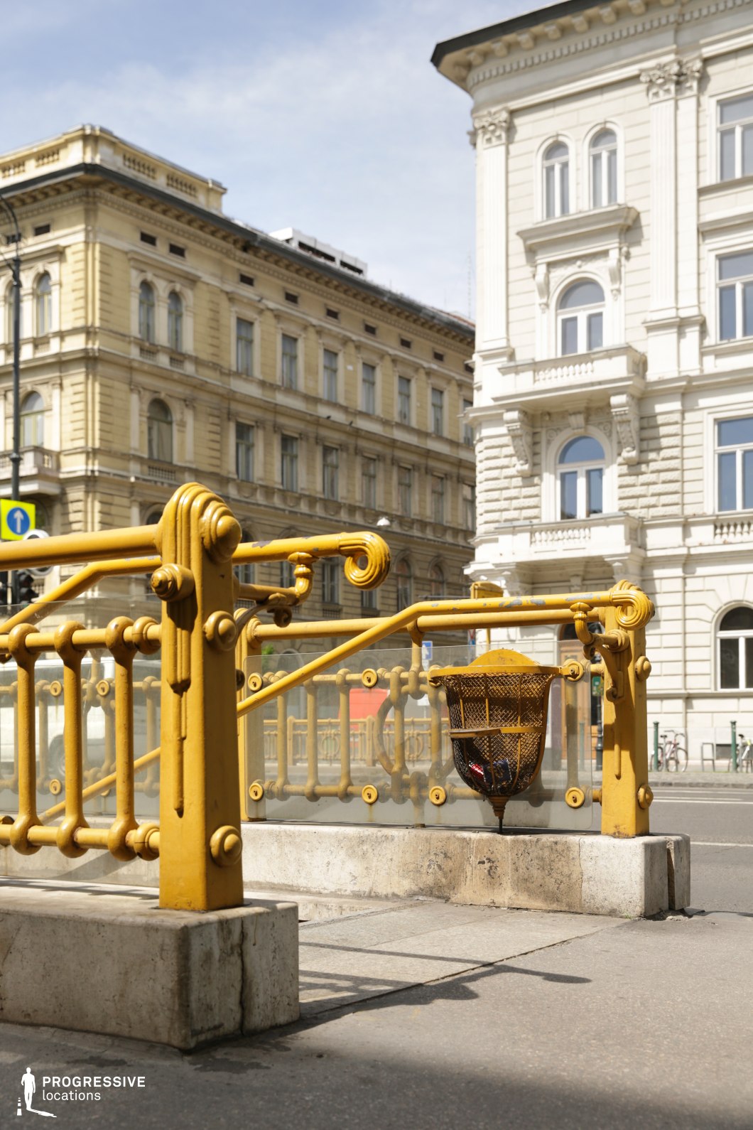 This image showcases an elegantly designed underground metro entrance with intricate yellow wrought-iron railings, featuring curved decorative elements and frosted glass panels. The stairway leads downward, framed by a matching metal trash receptacle integrated into the structure. In the background, grand neoclassical buildings with ornate facades, arched windows, and decorative cornices create a sophisticated European cityscape. The contrast between the detailed metalwork and the stately architecture enhances the location’s historical charm. This setting is ideal for filming period dramas, refined urban sequences, or classic European cityscapes, offering a visually rich and timeless backdrop.