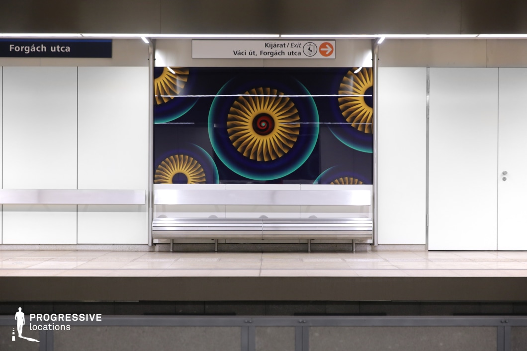 This image depicts a modern subway station platform with a minimalist design. A metallic bench is placed against a white wall, featuring a vibrant graphic design in the background. The graphic consists of swirling, abstract shapes in yellow and blue, creating a striking contrast against the clean, neutral tones of the surrounding architecture. The design elements, such as the smooth surface of the bench and the organized signage above, contribute to the contemporary, urban atmosphere. This location is well-suited for filming genres such as urban thrillers, sci-fi, or artistic dramas, offering a visually dynamic and modern backdrop that enhances both wide shots and artistic compositions.