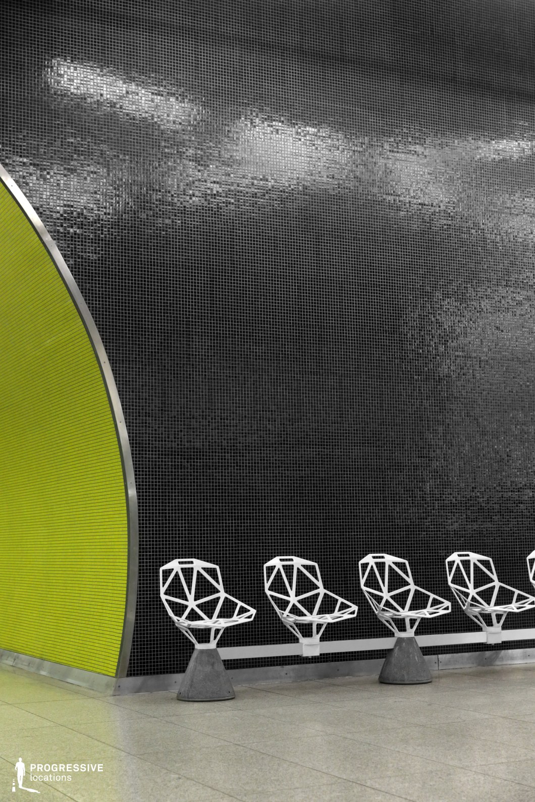 This modern subway station features geometric white chairs against a striking black tiled wall with a vibrant lime green section, creating a sharp contrast. The clean lines and angular shapes of the chairs, combined with the bold color scheme, give the space a contemporary, futuristic feel. This location is perfect for films in genres like science fiction, thriller, or drama, providing a visually dynamic and industrial backdrop for high-tech, urban scenes.