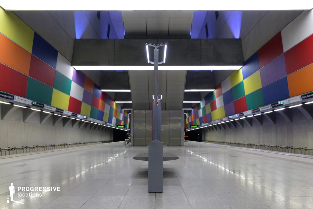 This metro station features a striking contrast between brutalist architecture and playful, multicolored geometric wall panels. The symmetrical composition is enhanced by sleek metallic lighting fixtures and large, suspended concrete beams that add depth to the space. The polished flooring reflects the ambient lighting, creating a bright and spacious atmosphere. The blend of bold colors with industrial materials results in a visually unique and modern aesthetic. This location is ideal for filming sci-fi, retro-futuristic, or artistic productions, offering a dynamic setting with a mix of urban minimalism and vibrant design elements.
