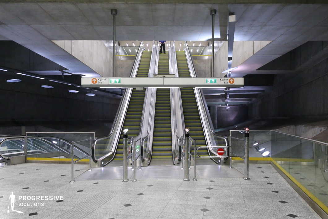 This escalator system is set within a brutalist-inspired underground structure, dominated by raw concrete surfaces and clean geometric lines. The interplay of artificial and natural light creates a dynamic contrast between the illuminated pathways and the shadowed recesses of the space. Overhead beams and angular ceiling structures add depth, while the glass railings and metallic finishes of the escalators provide a sleek industrial aesthetic. The monochromatic color palette and structured design make this an ideal location for futuristic, dystopian, or high-tech urban cinematography.