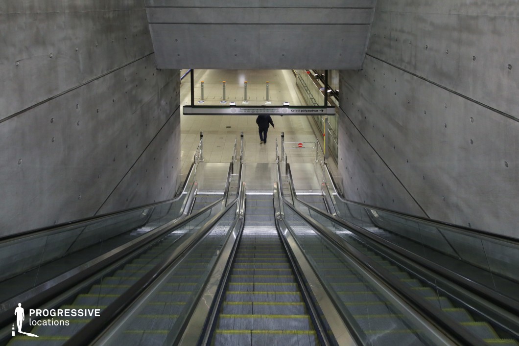 This escalator scene showcases a raw industrial aesthetic, with smooth, unadorned concrete walls punctuated by evenly spaced bolt impressions. The descending perspective emphasizes the depth of the space, leading to a transit platform below. The polished metal handrails and glass side panels reflect ambient light, contributing to a sleek, modern feel. The space is illuminated by a mix of natural and artificial lighting, casting soft shadows on the clean, geometric surfaces. This location is ideal for cinematographic sequences requiring a stark, futuristic, or dystopian setting.