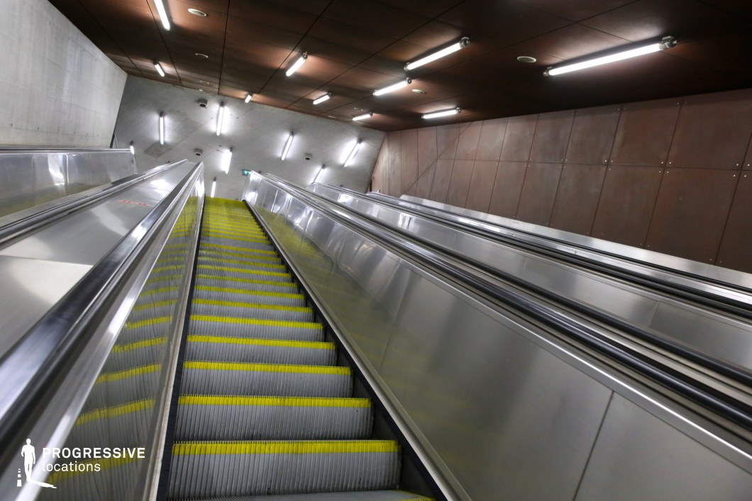 This metro station features a long, ascending escalator framed by sleek metallic railings and a mix of concrete and rust-toned wall panels. The overhead fluorescent lighting casts a cool, industrial glow, emphasizing the clean geometric lines and structured symmetry of the space. The contrast between the raw concrete and warm metal panels creates a futuristic yet utilitarian atmosphere. This location is ideal for filming sci-fi, dystopian, or thriller scenes, offering a striking urban setting with a sense of movement and architectural depth.