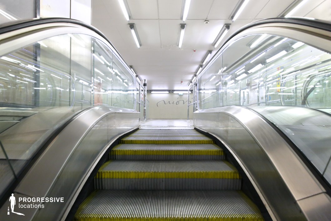 This image showcases a sleek, modern escalator with transparent glass walls and a brushed steel frame, highlighted by yellow-edged steps. The bright, even lighting and smooth, curved lines create a clean, industrial atmosphere, while the reflective surfaces and geometric shapes enhance the minimalist design. The space exudes a contemporary urban feel, making it an ideal location for filming action, thriller, or science fiction genres that require a modern, futuristic setting. Its spacious, dynamic layout offers versatile options for dramatic and motion-focused scenes.