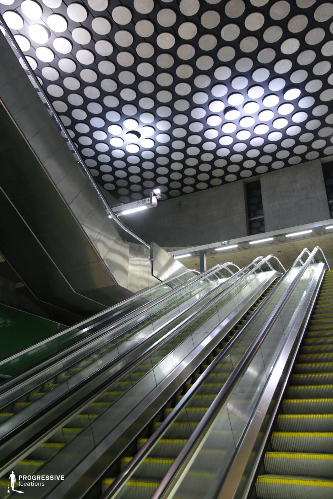 This escalator scene is defined by its striking geometric ceiling, featuring a grid of circular recesses with illuminated sections, creating a surreal and futuristic ambiance. The polished metal railings and reflective surfaces add depth, while the contrasting concrete and stainless steel architecture emphasize the industrial aesthetic. The escalators, with their sleek design and yellow-edged steps, lead the viewer's eye upwards, making this location ideal for sci-fi, high-tech, or dystopian cinematic settings.