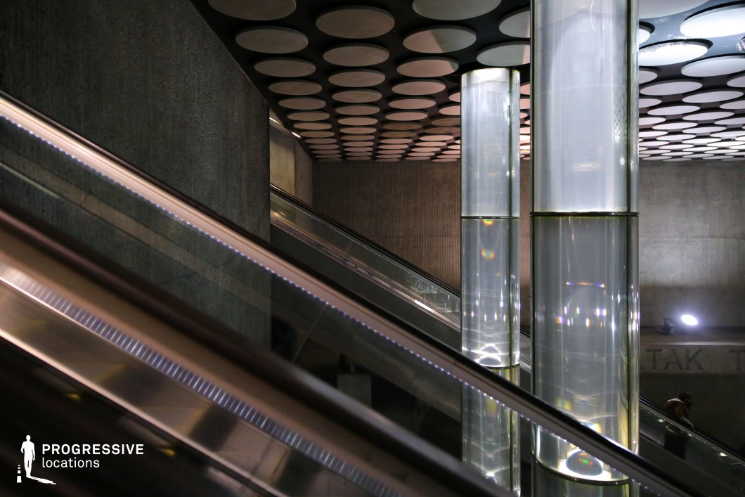 This metro station features a unique and futuristic design, highlighted by tall, cylindrical glass columns that emit soft, diffused light. The reflective surfaces of the escalators and railings enhance the modern aesthetic, while the dark, textured concrete walls create a striking contrast. The ceiling is adorned with a pattern of circular recessed panels, adding depth and rhythm to the architectural composition. The combination of sleek materials, geometric lighting, and industrial tones makes this location ideal for filming sci-fi, cyberpunk, or high-tech thriller scenes, offering a visually dynamic and immersive urban setting.