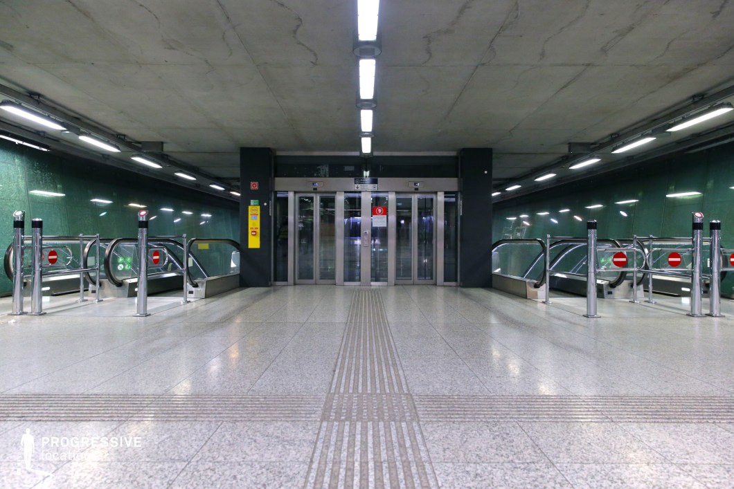 This image showcases a modern underground transportation hub with a sleek, industrial aesthetic. The space is defined by polished concrete ceilings, textured green glass walls, and a reflective tiled floor that enhances the ambient lighting. A centrally positioned elevator with metallic sliding doors serves as a focal point, flanked symmetrically by two sets of escalators descending into the station. Stainless steel barriers with red 
