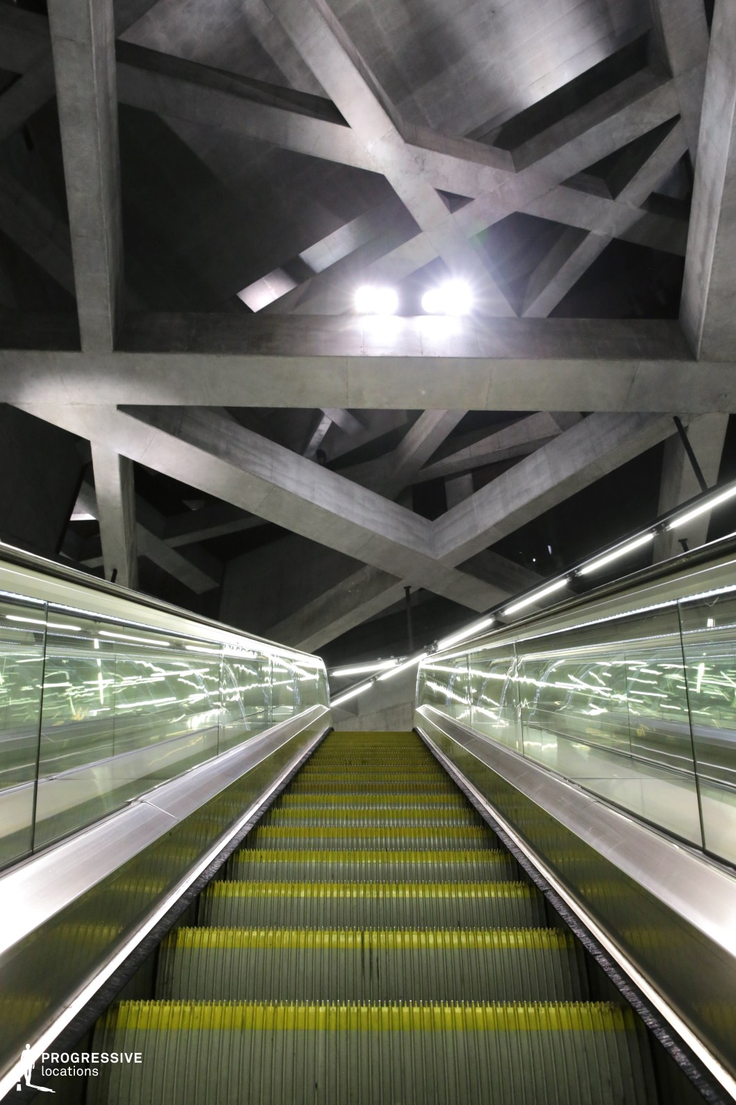 This metro station features a striking escalator passage framed by reflective glass railings and illuminated by sleek, linear lighting. The focal point is the ceiling’s intricate geometric network of raw concrete beams, forming a web-like structure that enhances the industrial, futuristic atmosphere. The perspective draws the eye upward, emphasizing scale and depth while the metallic surfaces reflect ambient light, creating a dynamic visual effect. The stark brutalist design, combined with the escalator’s symmetry and the dramatic lighting, makes this location ideal for filming sci-fi, dystopian, or cyberpunk scenes, evoking a high-tech, high-tension environment.