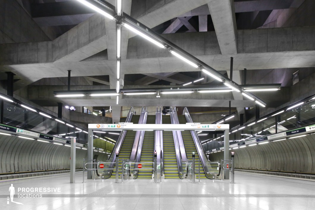 This modern subway station features a striking geometric design with a spacious concrete platform, highlighted by sharp angles and bright artificial lighting. The central escalators and clear directional signage emphasize the functional layout, while the industrial aesthetic, with smooth concrete and metal elements, creates a futuristic, minimalistic atmosphere. The open space and strong lines in the architecture enhance the modern, urban feel, making it ideal for urban, sci-fi, action, or thriller films.