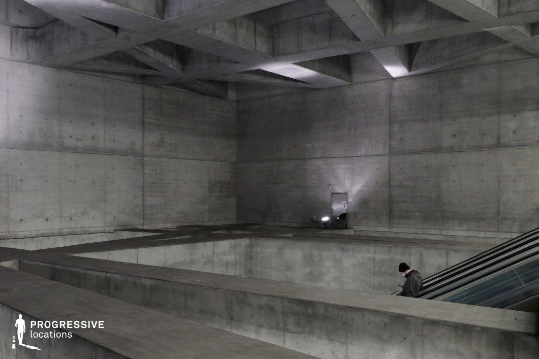 This vast, monolithic interior space is defined by its raw concrete surfaces, sharp geometric forms, and exposed structural framework. The intersecting beams in the ceiling create a dramatic pattern of light and shadow, emphasizing the industrial brutalist aesthetic. The stark, unadorned walls and deep recesses add a sense of depth and isolation, while the descending escalator integrates modern functionality into the otherwise austere design. This location is ideal for filming dystopian, sci-fi, or psychological thriller scenes, offering a cold, imposing, and futuristic setting that evokes themes of control, surveillance, or solitude.