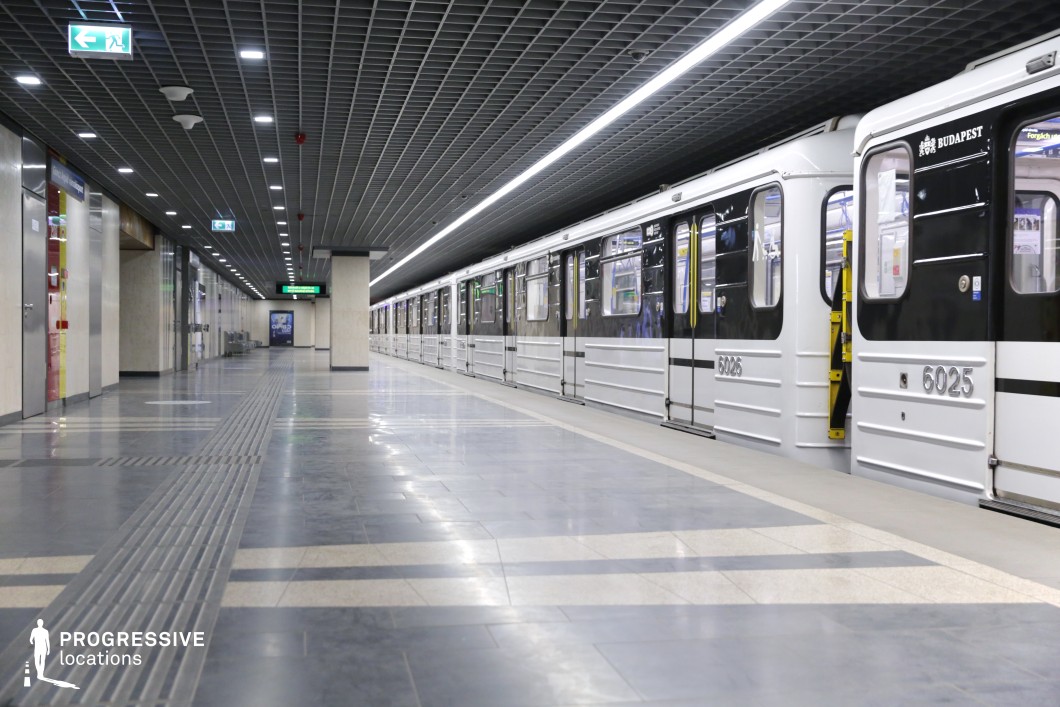 The image showcases a contemporary subway station with a sleek, industrial design, featuring a long, well-lit platform alongside black and silver subway cars. The space is defined by a grid-patterned ceiling, polished flooring with contrasting tones, and functional green directional signage. The modern, minimalistic materials, such as glossy tiles and metallic finishes, create an urban, efficient atmosphere. This versatile location is ideal for filming genres like action, thriller, or urban dramas, offering a spacious, clean backdrop that works well for both wide shots and dramatic scenes.