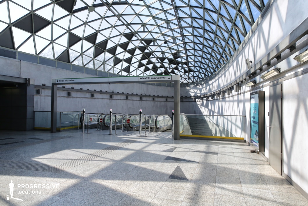 This contemporary transit station features a striking domed ceiling composed of a geometric glass framework, allowing ample natural light to filter through. The interplay of light and shadow creates dynamic patterns on the polished stone flooring, enhancing the architectural depth of the space. The curved walls and clean metallic accents contribute to the modern, futuristic aesthetic. The escalators, enclosed in transparent glass railings, add a sense of movement, making the location ideal for cinematic sequences emphasizing openness, transition, and futuristic design.