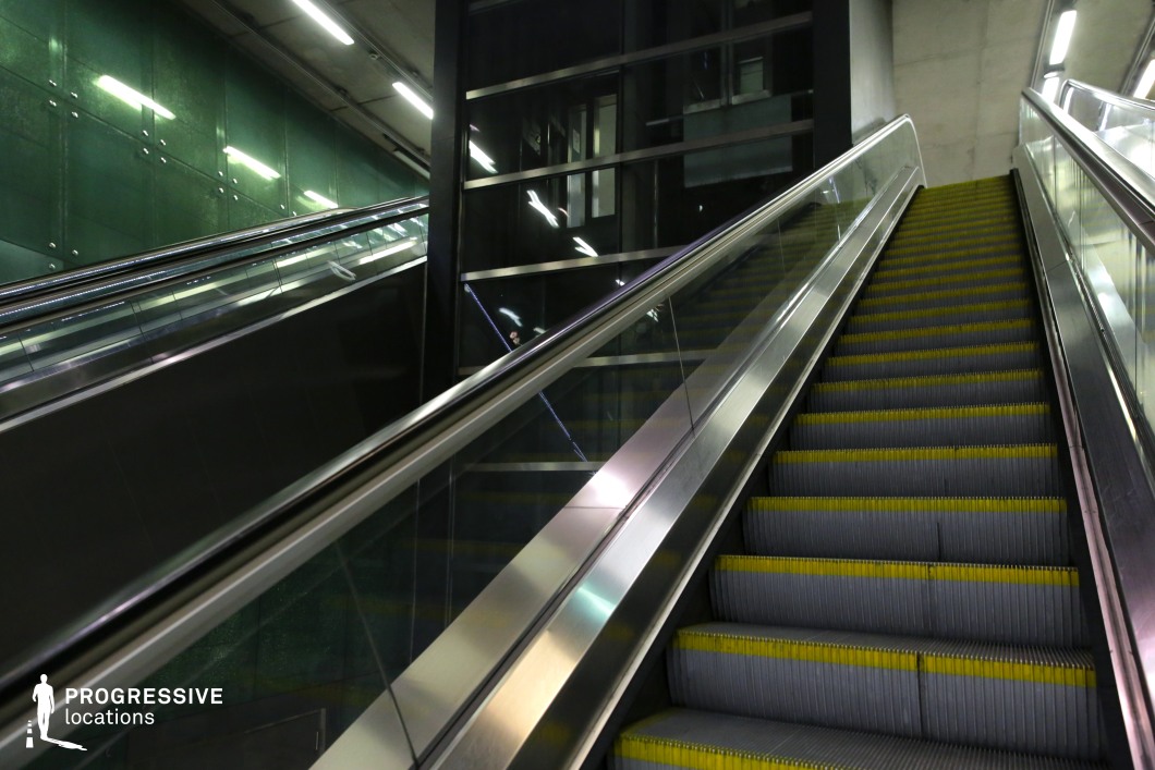 This image presents a modern escalator with a polished stainless steel and glass railing, ascending alongside a striking green glass wall. The reflective surfaces of the escalator and the glass panels contribute to a futuristic ambiance, enhanced by overhead strip lighting. The presence of an adjacent glass-paneled elevator structure adds a sense of depth and functionality. The combination of industrial materials and contemporary design makes this location ideal for urban-themed productions, sci-fi settings, or high-tech commercial visuals.