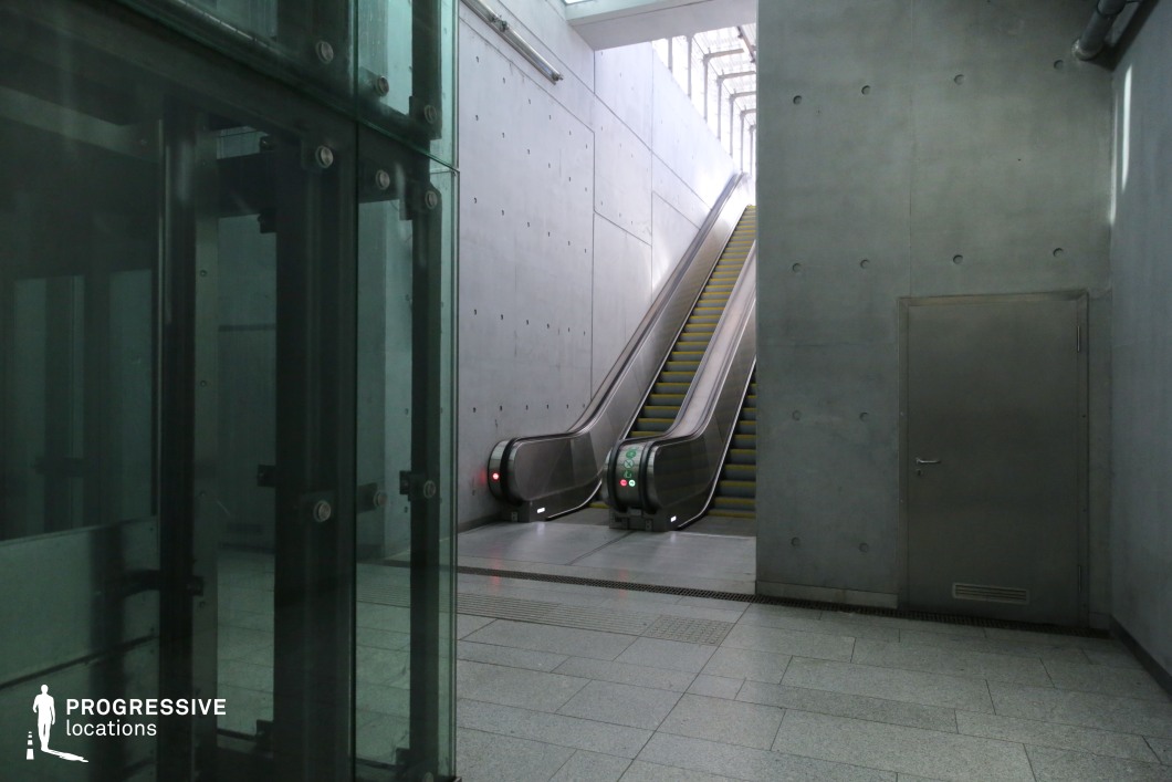This image features a sleek escalator set within a stark concrete environment, complemented by a glass-paneled elevator on the left. The industrial aesthetic is emphasized by exposed concrete walls, punctuated with a grid of recessed bolts, and a metallic door, adding to the space’s raw, utilitarian charm. Overhead natural light filters through a geometric skylight, casting soft illumination onto the scene. The combination of materials—glass, steel, and concrete—creates a futuristic and minimalist atmosphere, making this location ideal for high-tech, corporate, or dystopian cinematic settings.