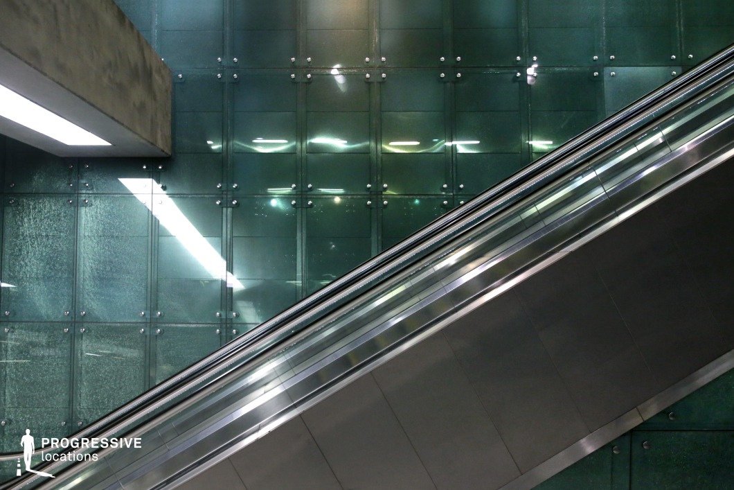 This image showcases a sleek escalator set against a striking green glass-paneled wall. The panels are structured with industrial metal fastenings, giving a contemporary and futuristic aesthetic. The interplay of reflections and diffused artificial lighting creates a dynamic visual effect. The contrast between the smooth metallic surfaces of the escalator and the textured glass panels adds depth to the scene. Ideal for film or commercial shoots requiring a modern, high-tech, or minimalist urban setting.
