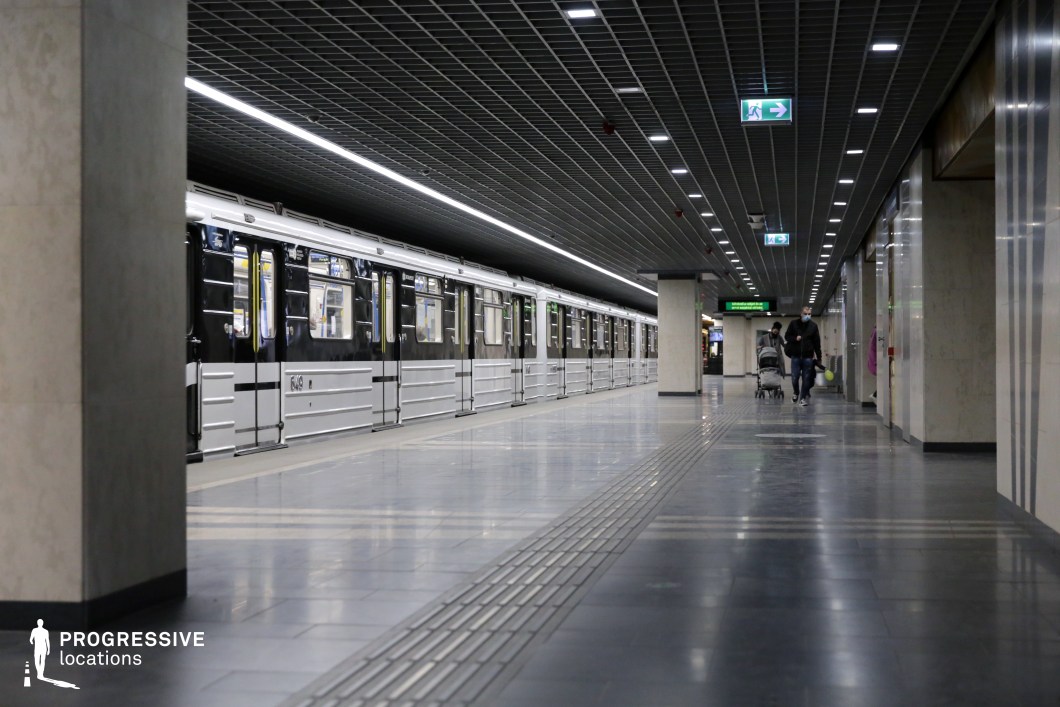 This image presents a modern subway platform with a sleek design, highlighted by a black and silver subway train running along the length of the platform. The space features a geometric grid ceiling with bright, evenly distributed lighting, which adds to the clean, industrial atmosphere. The smooth, polished tiles on the floor create a symmetrical pattern that complements the organized, minimalistic surroundings. The platform is spacious, with clear signage and wide, open walkways. This location is well-suited for filming in genres such as urban thrillers, action, or drama, offering a contemporary and visually dynamic backdrop that enhances a variety of scenes, from high-paced action sequences to more intimate, dramatic moments.
