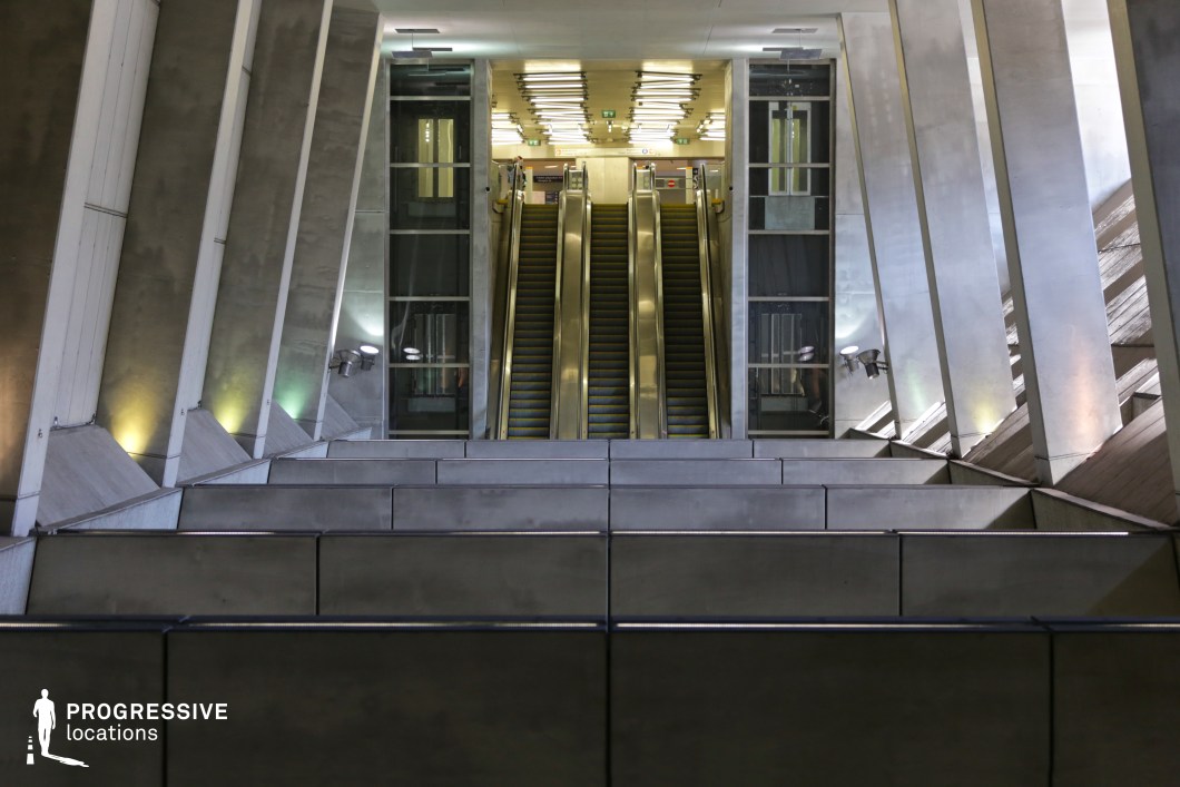 This grand transit station features a striking architectural design dominated by raw concrete and steel elements. The symmetrical layout of the escalators, framed by angled structural beams, creates a dramatic sense of depth and movement. Vertical glass windows allow natural light to filter in, complementing the artificial overhead illumination. The interplay of shadows and reflections enhances the futuristic and industrial aesthetic, making this space ideal for cinematic sequences requiring a modern, high-tech, or dystopian backdrop.