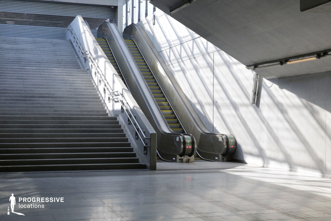 This image presents a striking urban transit space featuring a wide concrete staircase alongside two sleek escalators. The clean, geometric design is emphasized by sharp angles and smooth surfaces, creating a functional yet visually appealing setting. Natural light floods the area through a glass ceiling, casting dramatic shadows on the white walls and polished floor, enhancing the space’s modern aesthetic. The stainless steel handrails and escalator mechanisms contrast with the raw concrete, reinforcing the industrial yet contemporary atmosphere. This location is ideal for architectural cinematography, futuristic themes, or minimalist urban storytelling.