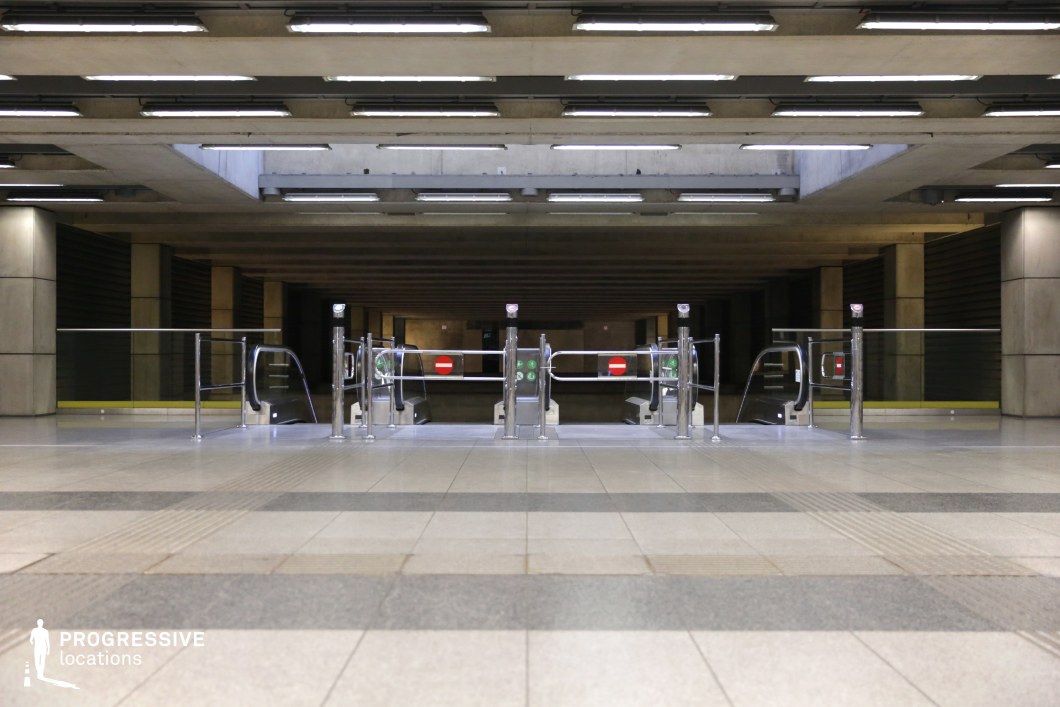 This underground passageway features a raw industrial aesthetic, dominated by exposed concrete beams and a repetitive linear lighting system. The structured ceiling pattern creates depth, while the sleek metallic turnstiles and glass railings contrast with the brutalist architectural elements. The combination of artificial lighting and the deep shadows in the background adds a futuristic or dystopian ambiance. The expansive floor space, marked with geometric tile patterns, enhances the location’s versatility for cinematic and artistic compositions.