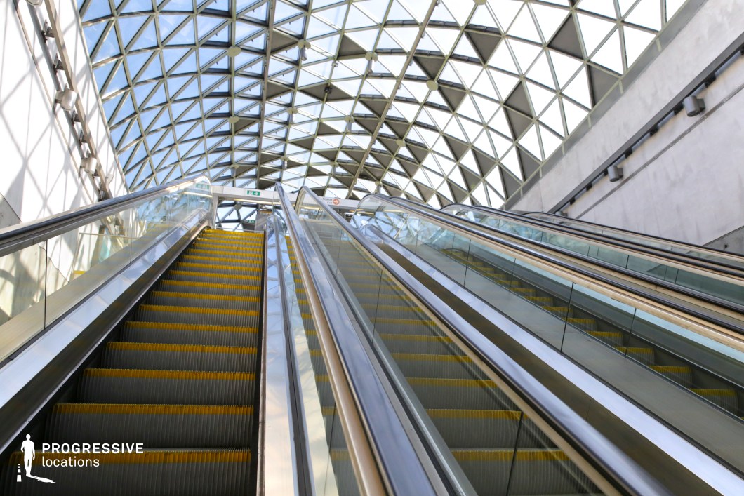 This modern transit station features a striking geometric glass ceiling, allowing an abundance of natural light to flood the space. The escalators, framed by sleek glass and steel railings, lead upwards, emphasizing movement and openness. The interplay of light and shadow created by the intricate ceiling structure adds a dynamic visual element, making the space ideal for futuristic, corporate, or high-tech cinematic settings. The combination of raw concrete, metallic surfaces, and transparent glass enhances the contemporary architectural appeal.