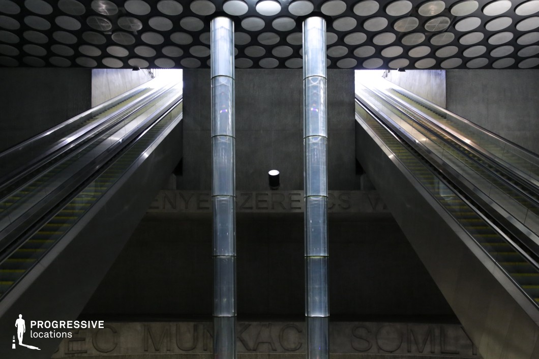 This metro station features a dramatic and futuristic design, with towering glass light columns rising between two symmetrical escalators. The textured concrete walls and circular-patterned ceiling create a bold industrial aesthetic, while the openings above allow natural light to stream in, adding depth and contrast. The metallic escalators reflect the ambient light, enhancing the sleek, high-tech atmosphere. The stark geometry and interplay of light and shadow make this location ideal for filming sci-fi, dystopian, or cyberpunk scenes, offering a visually striking and immersive urban environment with a sense of scale and grandeur.