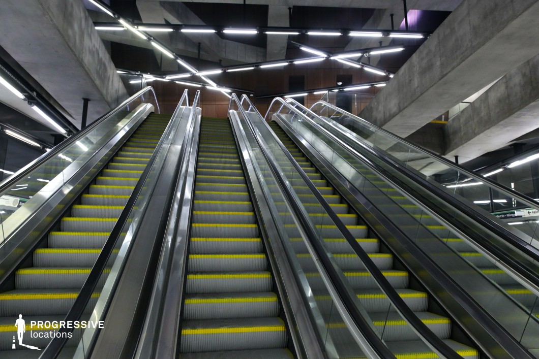 This metro station features an imposing set of escalators framed by sleek glass and metallic railings, leading upward into a structured brutalist space. The raw concrete beams contrast sharply with the dynamic arrangement of neon-style overhead lights, creating an intricate geometric pattern that enhances the futuristic aesthetic. The perspective emphasizes depth and movement, making the location feel vast and cinematic. The cold industrial tones, sharp angles, and dramatic lighting make this an ideal setting for cyberpunk, dystopian, sci-fi, or high-tech thriller productions, offering a visually striking and immersive urban environment.