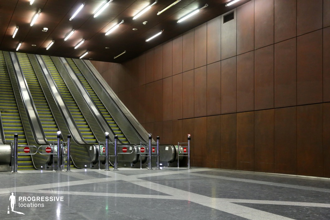 This metro station features an imposing set of escalators framed by large, rust-colored metal panels that add warmth and depth to the industrial design. The sleek metallic railings and dark, polished flooring create a strong visual contrast, while the overhead fluorescent lighting enhances the stark, modern aesthetic. The geometric floor pattern and controlled symmetry contribute to a structured, high-tech atmosphere. This location is ideal for filming dystopian, sci-fi, or cyberpunk scenes, offering a dramatic urban setting that evokes a futuristic yet industrial ambiance.