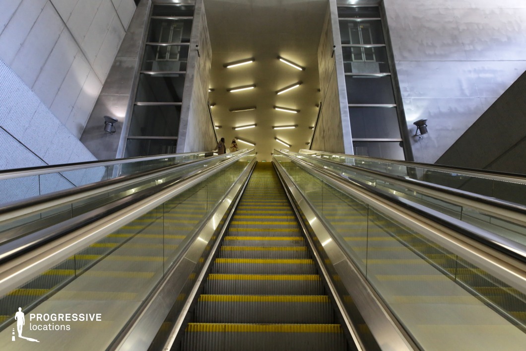 This metro station features a long, imposing escalator framed by sleek glass railings and reflective metallic surfaces, leading toward a luminous upper level. The geometric arrangement of neon-style overhead lights creates a dynamic visual rhythm, reinforcing the station’s futuristic and high-tech aesthetic. The combination of smooth concrete walls, industrial lighting, and sharp architectural lines adds depth and grandeur to the space. The minimalistic yet monumental design makes this location ideal for filming sci-fi, dystopian, or cyberpunk scenes, offering a sleek, high-tech setting with a cinematic sense of movement and scale.