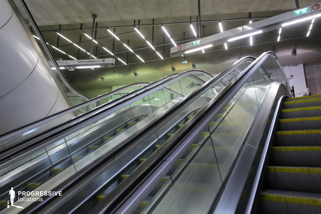 This modern transit station features a sleek escalator system with reflective metal handrails and glass panels, creating a futuristic and industrial aesthetic. The ceiling is adorned with a geometric grid of linear LED lights, providing even illumination and accentuating the clean architectural lines. Exposed concrete elements and subtle green lighting contribute to the contemporary, high-tech atmosphere. The structure's design, incorporating smooth metallic curves and sharp angles, makes this location an excellent setting for urban sci-fi, cyberpunk, or minimalist architectural photography.