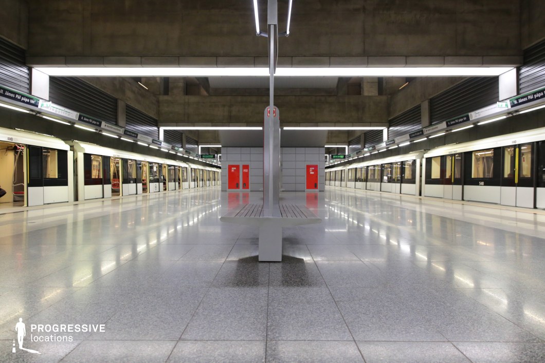 This metro station showcases a bold brutalist design with raw concrete beams, industrial lighting, and a perfectly symmetrical composition. The platform is framed by sleek, modern metro trains on both sides, while the polished tiled flooring reflects the overhead lighting, enhancing the sense of depth and order. The minimalistic bench at the center, along with red accent doors in the background, adds contrast to the neutral tones. The combination of geometric precision, industrial materials, and an open, spacious layout makes this location ideal for filming sci-fi, dystopian, or thriller scenes, providing a stark yet futuristic urban setting.