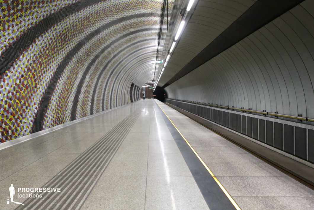 This striking metro platform features a curved tunnel design, with one side adorned by an intricate mosaic tile pattern in shades of yellow, red, and black. The opposite wall presents a smooth, metallic finish with arched paneling, creating a contrasting yet harmonious blend of textures. Overhead lighting runs parallel to the tracks, casting a soft, even glow. The polished stone flooring enhances the station’s modern, reflective aesthetic. The tunnel’s receding perspective adds depth, making it an ideal location for dynamic cinematography, futuristic settings, or high-end fashion shoots.