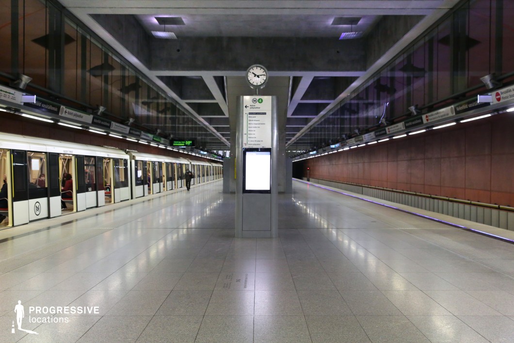 This metro platform features a striking blend of industrial and modern architectural elements, with exposed concrete beams and polished floors reflecting the overhead lighting. The symmetrical design is enhanced by a central digital display and a clock, creating a focal point in the station. The contrasting materials—warm rust-colored wall panels and sleek metallic fixtures—add depth to the setting. A waiting train with illuminated interiors adds a dynamic element, making this location ideal for urban transit-themed productions, futuristic settings, or commercial shoots requiring a structured, high-tech aesthetic.