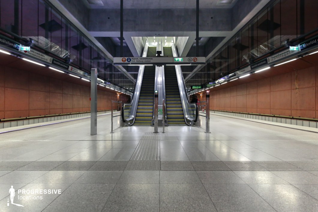 This metro station showcases a symmetrical, modern-industrial design, with a pair of escalators centrally positioned beneath a geometric concrete ceiling. The walls feature large, rust-colored panels that contrast with the sleek metallic and glass elements, adding depth and texture to the space. The polished tiled floor reflects the artificial lighting, enhancing the station’s clean and structured aesthetic. The combination of raw concrete, warm-toned walls, and sharp architectural lines creates a balance between functionality and visual appeal. This location is ideal for filming thriller, dystopian, or cyberpunk-themed scenes, offering a structured yet slightly gritty urban setting.