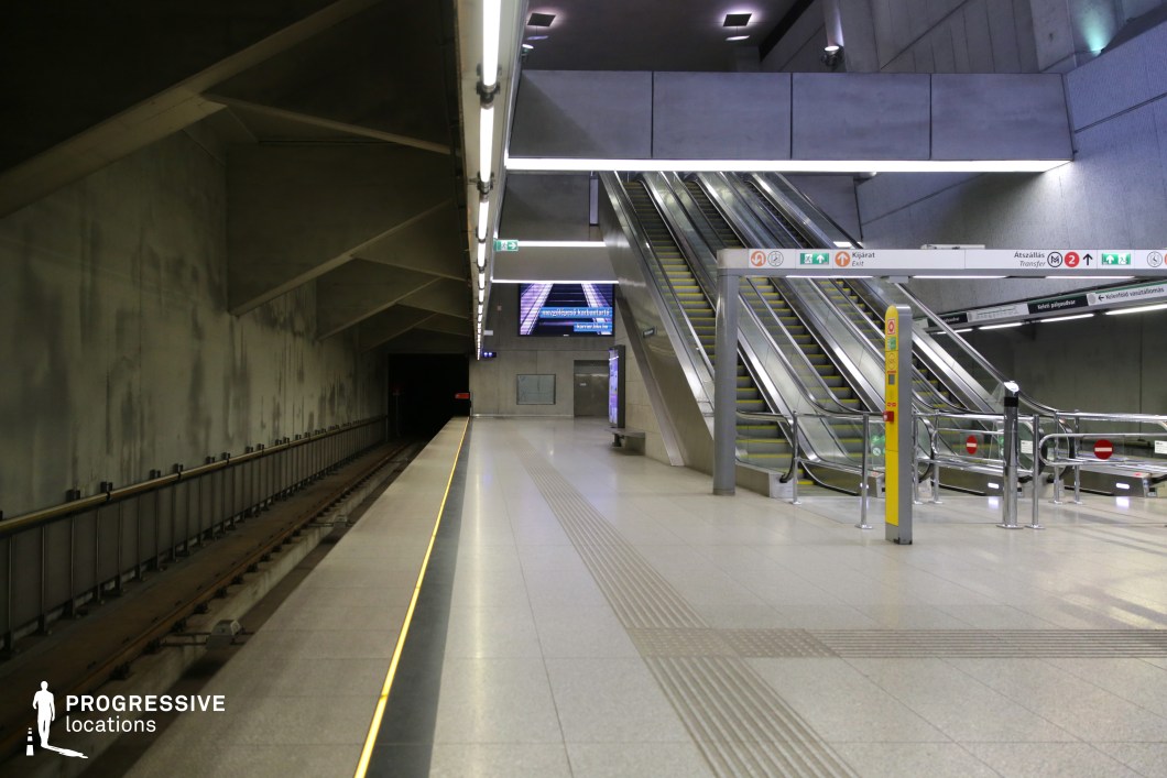 This underground transit platform showcases a sleek, industrial aesthetic with raw concrete walls and geometric ceiling supports. The escalators, framed by metallic railings and illuminated signage, create a structured, directional flow, guiding passengers toward the upper levels. The dark tunnel entrance on the left adds depth and contrast, emphasizing the station's vastness. The clean, minimalist design and bright linear lighting make this location ideal for futuristic, high-tech, or dystopian film settings.
