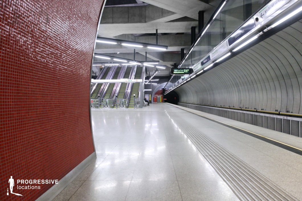 This image features a contemporary subway platform with curved metallic walls and a vibrant red tiled accent, complemented by bright linear lighting along the ceiling. The minimalistic design, with smooth flooring and clean geometric lines, creates a striking contrast between the red wall and the industrial gray tones of the tunnel and escalators. This location is ideal for modern, futuristic, or sci-fi films, offering a visually dynamic backdrop for sleek, high-tech scenes.