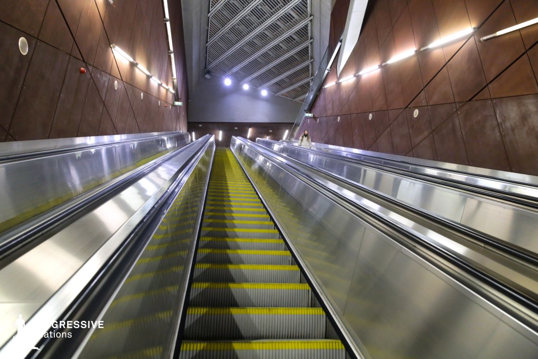 This metro station features a monumental escalator system encased by rust-toned metal panels with a geometric pattern, adding depth and texture to the industrial design. The metallic surfaces of the escalators reflect the ambient lighting, creating a sleek, futuristic atmosphere. The ceiling structure, with its slanted grid-like panels and integrated spotlights, enhances the sense of movement and scale. The combination of raw industrial materials and modern lighting makes this location ideal for filming dystopian, sci-fi, or cyberpunk scenes, offering a dramatic and high-tech urban setting with a bold architectural presence.