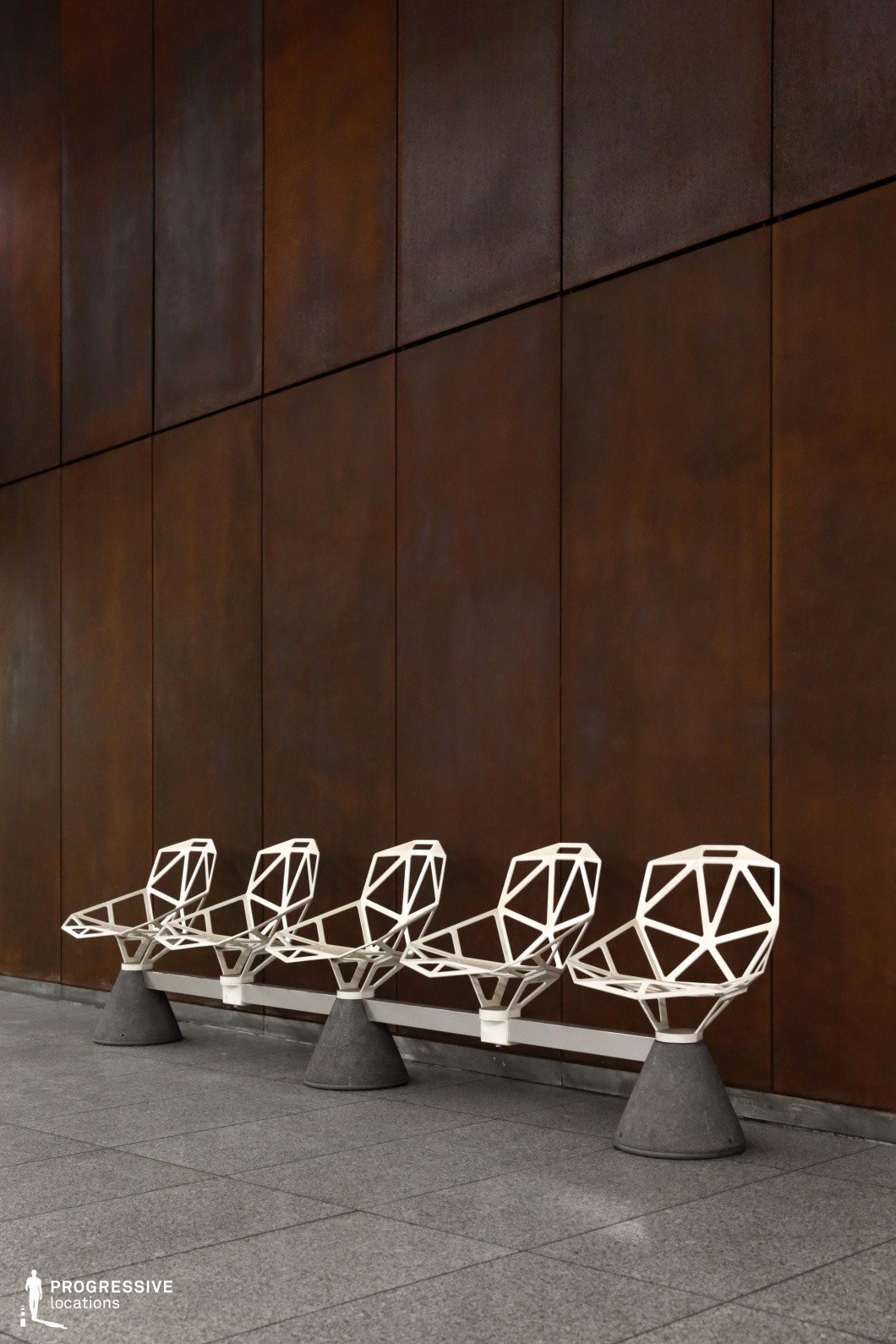 This metro station features a row of striking geometric metal chairs with a minimalist, futuristic design, set against a backdrop of large rust-toned wall panels. The contrast between the angular white metal framework of the seating and the warm, industrial surface of the wall creates a visually compelling composition. The polished concrete flooring adds to the structured, urban aesthetic, while the clean lines and repetition of elements emphasize modernity. This location is ideal for filming sci-fi, dystopian, or conceptual art productions, offering a refined yet industrial setting with a bold architectural presence.