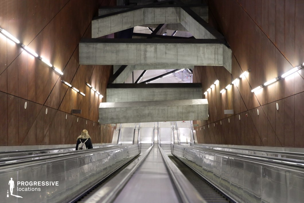 This large-scale escalator system is set within a striking architectural space that combines raw concrete elements with rust-toned paneling. The structural beams form an intricate geometric framework above, creating a sense of depth and complexity. Linear lighting along the walls emphasizes the sharp angles and industrial aesthetic, while the reflective metal surfaces of the escalators contrast with the matte, weathered textures of the walls. This location offers a dramatic, almost dystopian feel, making it an ideal backdrop for cinematic scenes requiring a futuristic, industrial, or psychologically intense atmosphere.