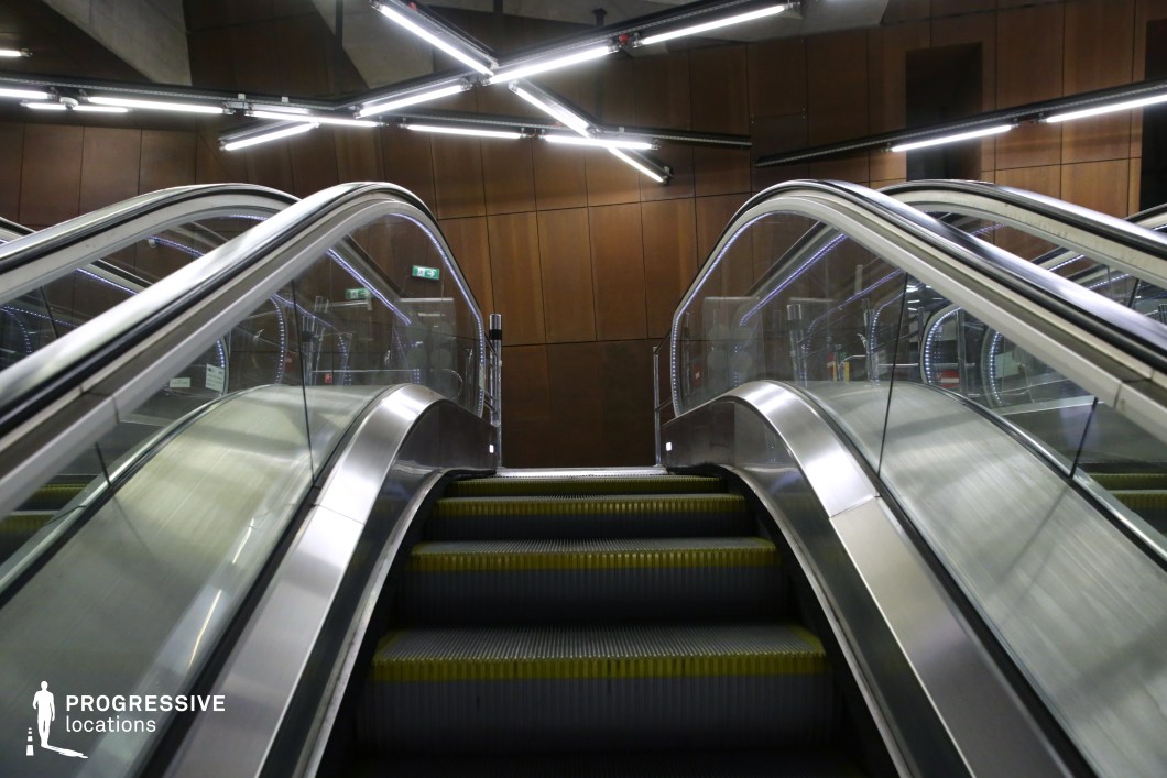 This metro station features a sleek, modern escalator system framed by curved glass railings and polished metallic surfaces. The rust-toned wall panels provide a striking contrast to the industrial design, while the angular, neon-style lighting fixtures above create a dynamic and futuristic atmosphere. The structured symmetry and reflective materials enhance the depth and visual complexity of the space. This location is ideal for filming sci-fi, cyberpunk, or action-thriller scenes, offering a sophisticated yet industrial backdrop with a high-tech urban aesthetic.