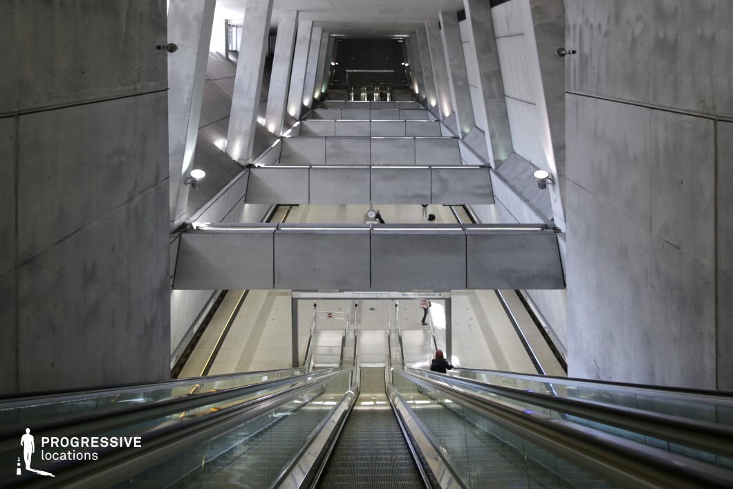 This metro station features an imposing, tunnel-like structure defined by raw concrete surfaces, angular support beams, and a cascading escalator system. The stark, industrial aesthetic is enhanced by the symmetrical layout and linear lighting, casting subtle shadows that emphasize depth and geometric precision. The reflective glass railings contrast with the rough concrete, adding a sleek, futuristic touch. Overhead crossbeams create a sense of enclosure, reinforcing the station’s monumental scale. This location is ideal for filming dystopian, sci-fi, or psychological thriller scenes, offering a visually striking and immersive urban setting with a cold, high-tech ambiance.