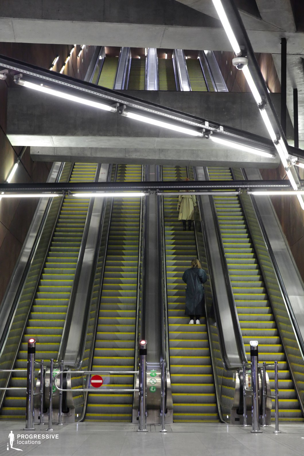 This metro station features a towering escalator system extending across multiple levels, framed by raw concrete beams and sharp geometric lighting fixtures. The exposed industrial ceiling, with its angular neon-style lights, adds a futuristic, high-tech atmosphere, while the sleek metallic railings and structured symmetry enhance the visual depth. The vast vertical scale and repetitive patterns of the escalators create a sense of movement and grandeur. This location is ideal for filming sci-fi, dystopian, or cyberpunk scenes, offering a visually striking setting with a modern, industrial aesthetic and an almost labyrinthine quality.