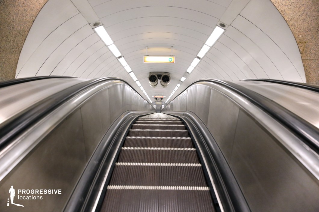 This image features a sleek, metallic escalator ascending in a minimalist subway station with smooth white walls and ceiling. The polished metal contrasts with the clean, modern design, enhanced by bright, even lighting that creates a futuristic, high-tech atmosphere. The symmetrical lines and simplicity of the space make it ideal for futuristic, sci-fi, or action films, offering a versatile backdrop for dynamic, urban-themed scenes.