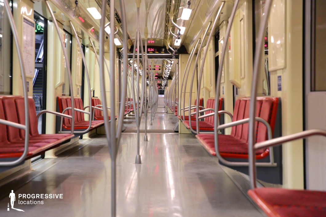 This metro train interior features a clean and symmetrical design with red cushioned seats and curved metallic handrails that create a dynamic visual rhythm. The warm yellow lighting complements the polished metal flooring, enhancing the sleek, contemporary aesthetic. Overhead digital displays provide real-time travel information, reinforcing the efficiency of this urban transit environment. The reflective surfaces and linear perspective make this space ideal for cinematic sequences, futuristic transport scenes, or advertising campaigns emphasizing modern mobility and urban connectivity.