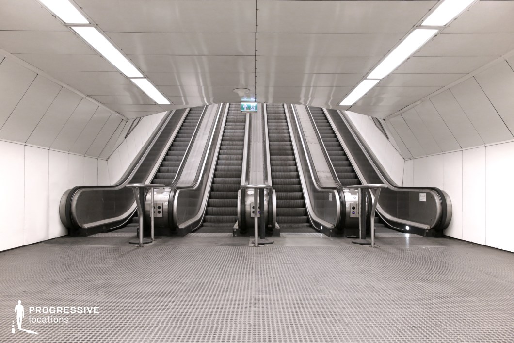 This image features a clean, minimalist subway space with two escalators framed by smooth metallic structures against a bright white background. The symmetrical composition, even lighting, and textured floor enhance the modern, industrial aesthetic, emphasizing the space’s functional simplicity. The sleek, futuristic vibe makes this location ideal for futuristic, action, or thriller films, offering versatility for dynamic, visually striking scenes.