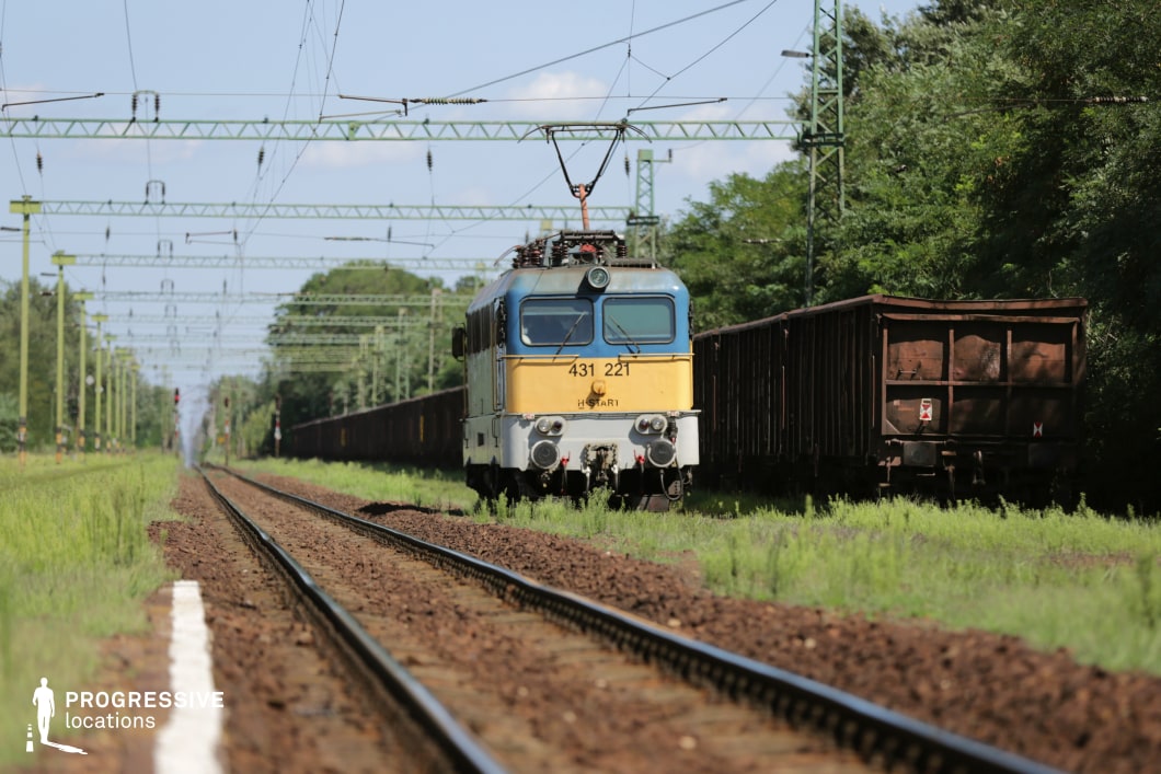 This low-angle shot captures a freight train moving along an electrified railway, emphasizing its imposing industrial presence. The locomotive, with its worn yellow and blue exterior, pulls a series of rusted cargo wagons, reinforcing a sense of rugged functionality. Overhead, a dense network of green metal electrical structures and power lines creates a repetitive pattern, adding depth and structure to the scene. The railway tracks stretch into the distance, lined with overgrown grass and trees, providing a contrast between industry and nature. This location is ideal for filming transportation sequences, industrial-themed narratives, or atmospheric travel montages, making it well-suited for drama, thriller, or documentary genres.