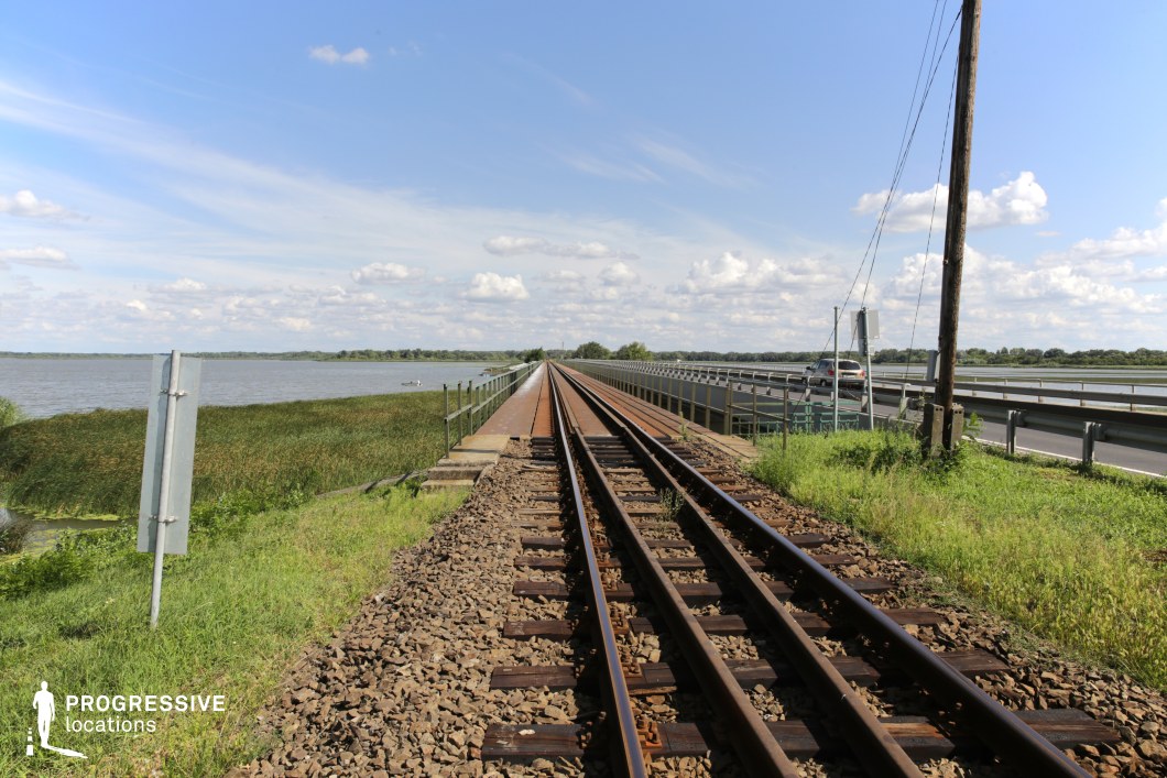This railway bridge extends in a perfectly straight line over a vast body of water, creating a strong sense of depth and perspective. The tracks are flanked by a parallel roadway, separated by a metal guardrail, reinforcing the transportation-focused setting. On the left, tall reeds grow along the water's edge, blending the infrastructure with the surrounding natural environment. The open sky and distant treeline contribute to a spacious, almost isolated atmosphere. This location is ideal for filming travel sequences, contemplative journeys, or dramatic escape scenes, making it well-suited for adventure, thriller, or road-movie genres.
