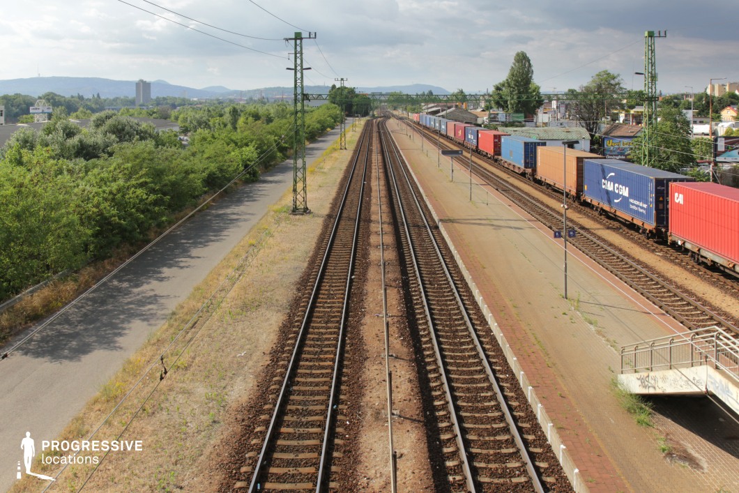 This wide-angle view of a railway corridor features multiple parallel train tracks stretching into the distance, emphasizing depth and perspective. A long freight train, loaded with colorful shipping containers, runs along the right side, adding an industrial and logistical character to the scene. The tracks are bordered by a paved platform and an overgrown service road, while tall green electrical poles with overhead wires enhance the structured, industrial aesthetic. In the background, a city skyline with low-rise buildings and distant hills provides a contrasting element of urbanization against the raw infrastructure. This location is ideal for filming transport-based sequences, industrial settings, or travel montages, making it suitable for action, drama, or documentary-style productions.