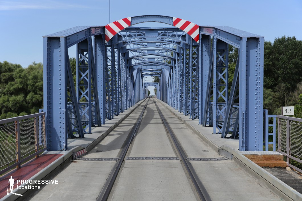 This striking blue steel truss bridge stretches across a river, its repeating metal framework creating a visually captivating tunnel-like effect. The structure is reinforced with riveted beams and latticework, giving it an industrial yet elegant aesthetic. The railway tracks run along a concrete pathway, with protective railings on either side, ensuring both functionality and safety. Red-and-white striped warning signs add a touch of contrast to the cool-toned metal. Surrounded by lush greenery, this location offers a dynamic setting for filming action sequences, travel montages, or dramatic crossings, making it ideal for adventure, thriller, or road-movie-style storytelling.
