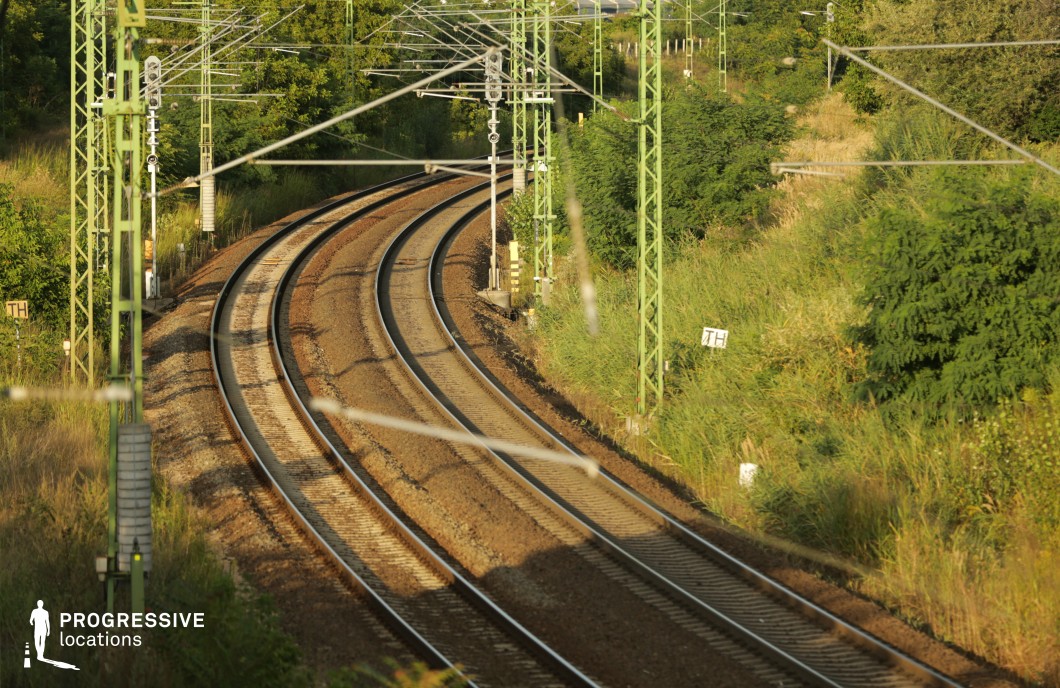 This railway scene features a set of curved tracks cutting through a landscape of wild grasses and dense green shrubs. Tall, green-painted metal poles support an intricate network of overhead power lines, creating a repeating pattern that adds depth and structure to the composition. The tracks, made of steel and embedded in gravel, reflect light, emphasizing the curvature and leading the eye into the distance. The surrounding vegetation gives the location a natural, slightly overgrown feel, contrasting with the industrial elements. This setting is ideal for filming travel sequences, reflective or suspenseful moments, and atmospheric shots suited for road movies, thrillers, or dramas centered around movement and transition.